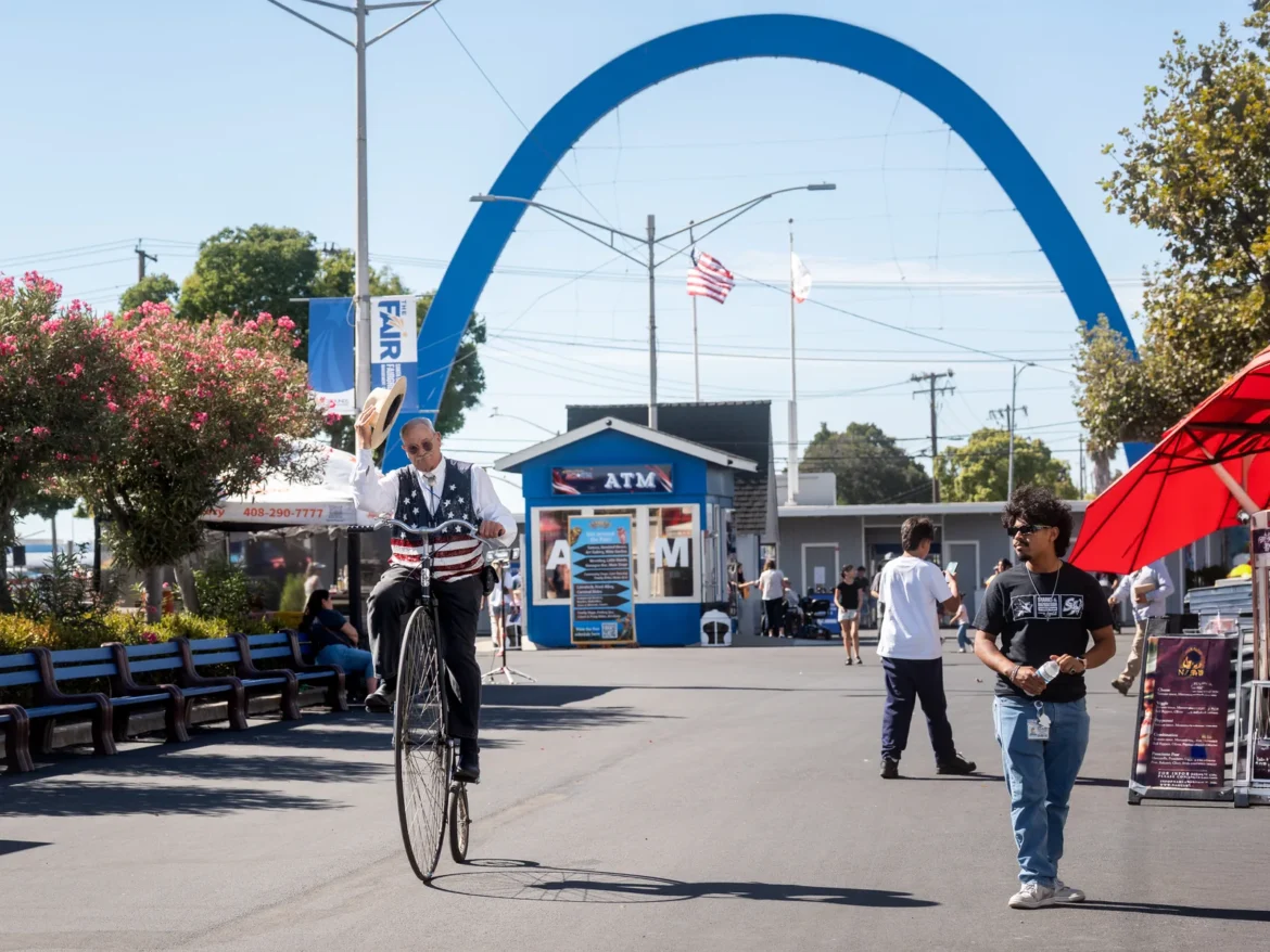 A unicyclist rides past the blue Fair entrance arch on a sunny afternoon.