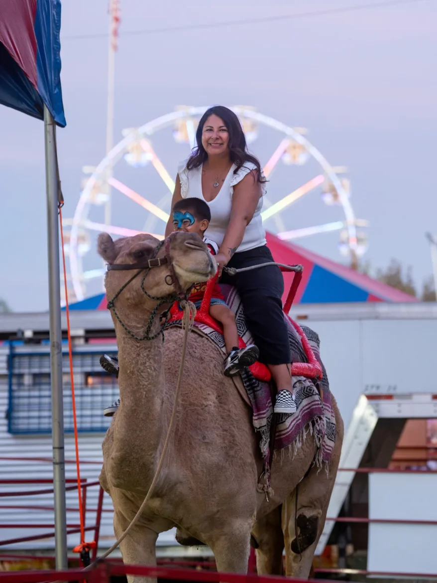 A mother and child ride a camel with the Ferris wheel behind them.